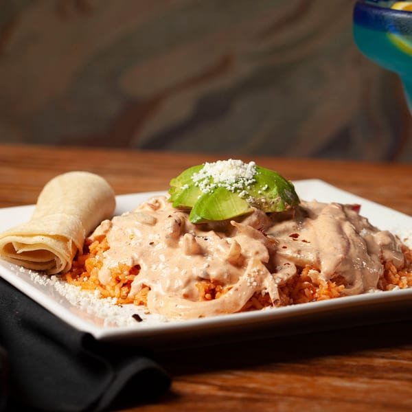 Mexican dish with chicken in creamy sauce, rice, avocado, and a tortilla on a white plate.
