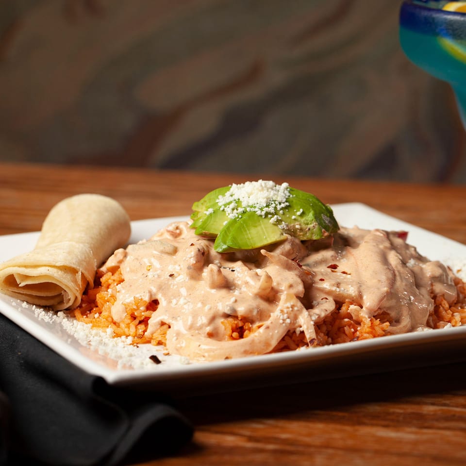 Mexican dish with chicken in creamy sauce, rice, avocado, and a tortilla on a white plate.