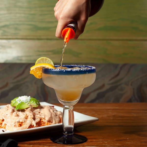 A hand pours liquid into a salt-rimmed margarita glass with citrus, next to a Mexican dish.