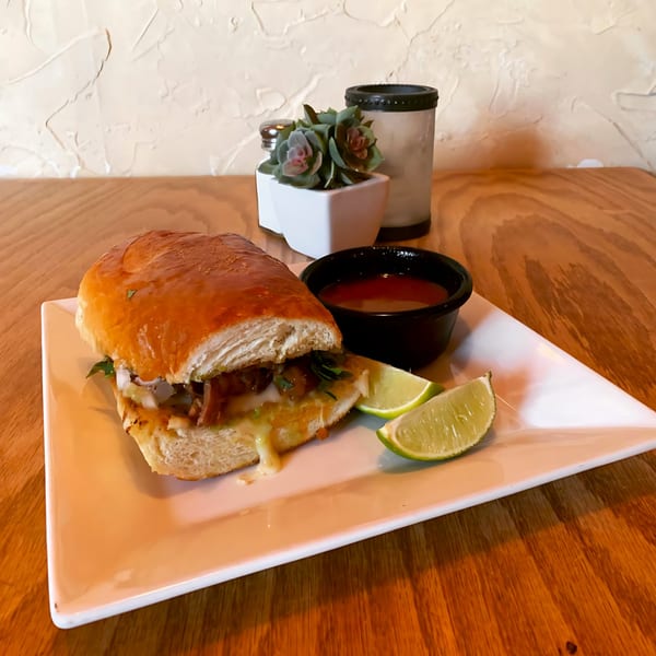 A Mexican torta sandwich with melted cheese, onions, and cilantro, served with lime wedges and salsa.