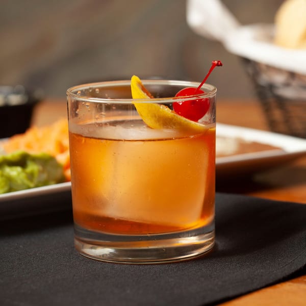 A glass of amber-colored drink with ice, a lemon slice, and a cherry on a black napkin.
