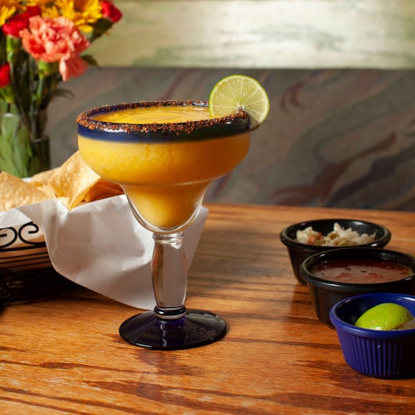 Mango margarita with a chili-lime rim and lime wedge, alongside chips and salsa on a wooden table.