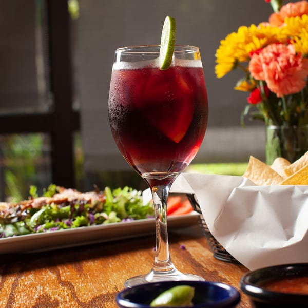A glass of red sangria with a lime wedge, salad, and tortilla chips on a wooden table.