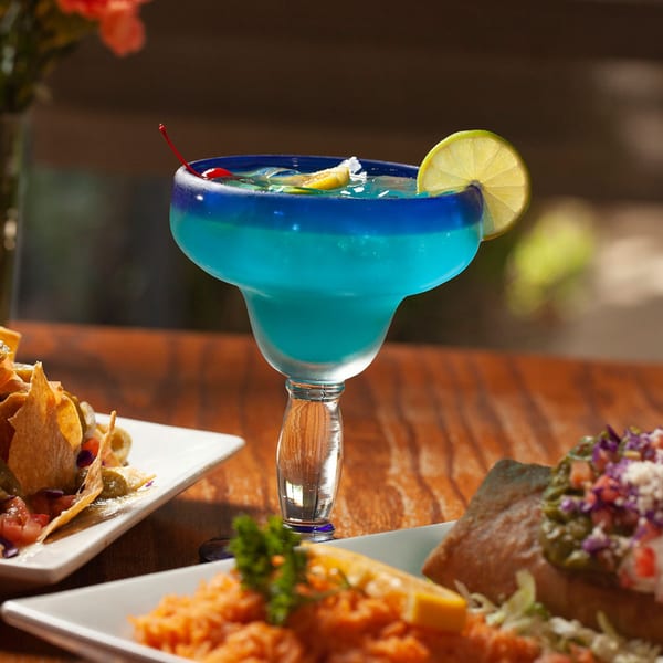 A blue margarita with a lime and cherry, surrounded by Mexican dishes on a wooden table.