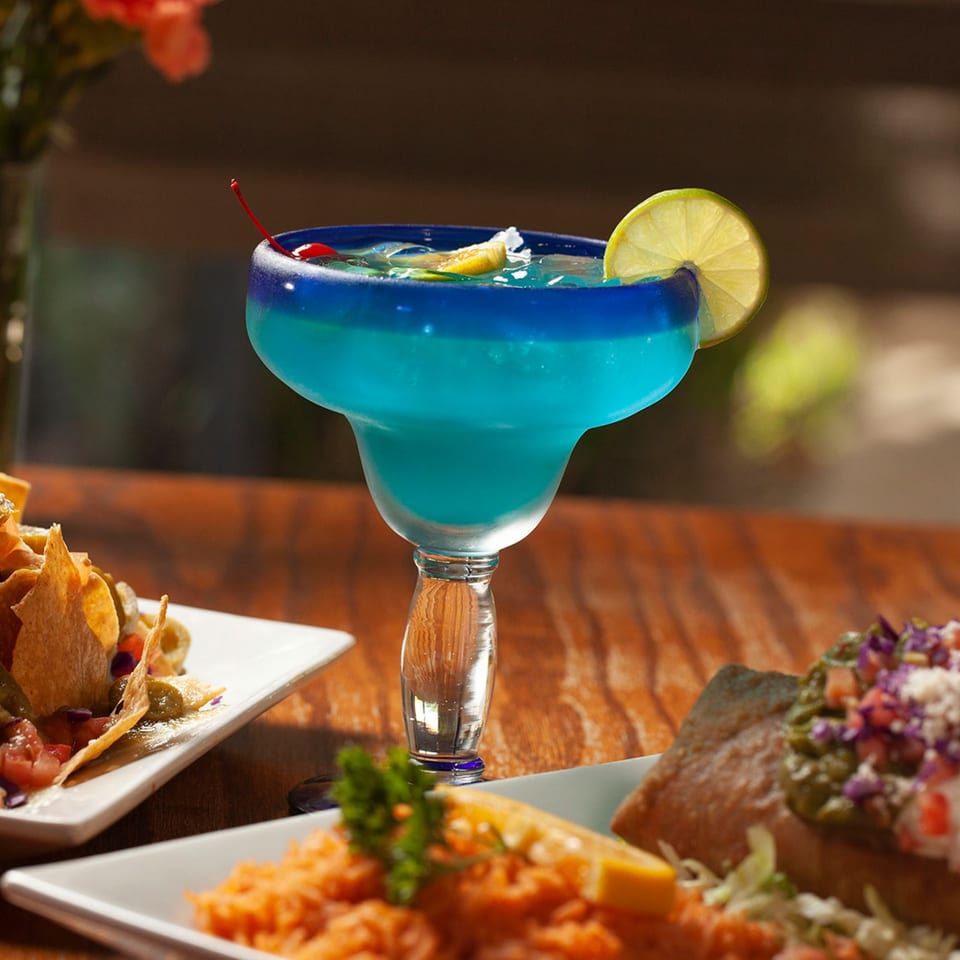 A blue margarita with a lime and cherry, surrounded by Mexican dishes on a wooden table.